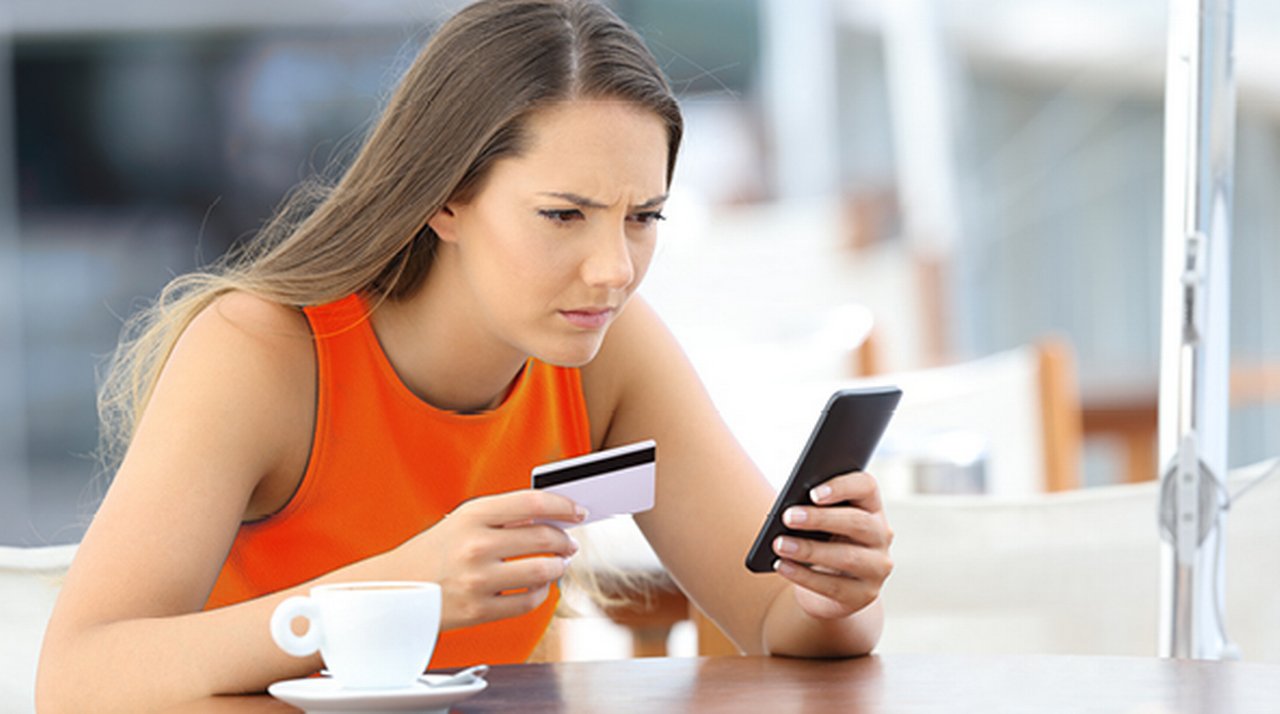 Frau an Kaffeetisch schaut angestrengt auf ihr Smartphone in der einen Hand, während sie eine Bankkarte in der anderen hält.