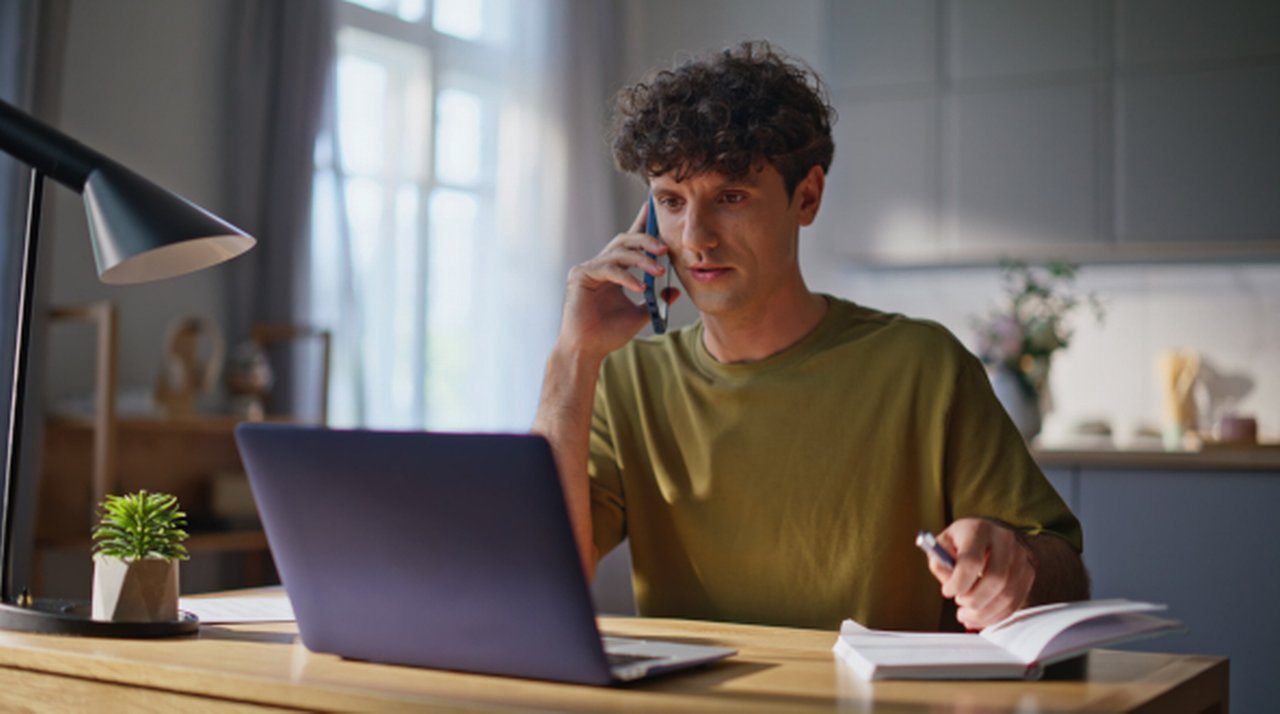 A man sits at the kitchen table on the phone, looking at his open laptop.