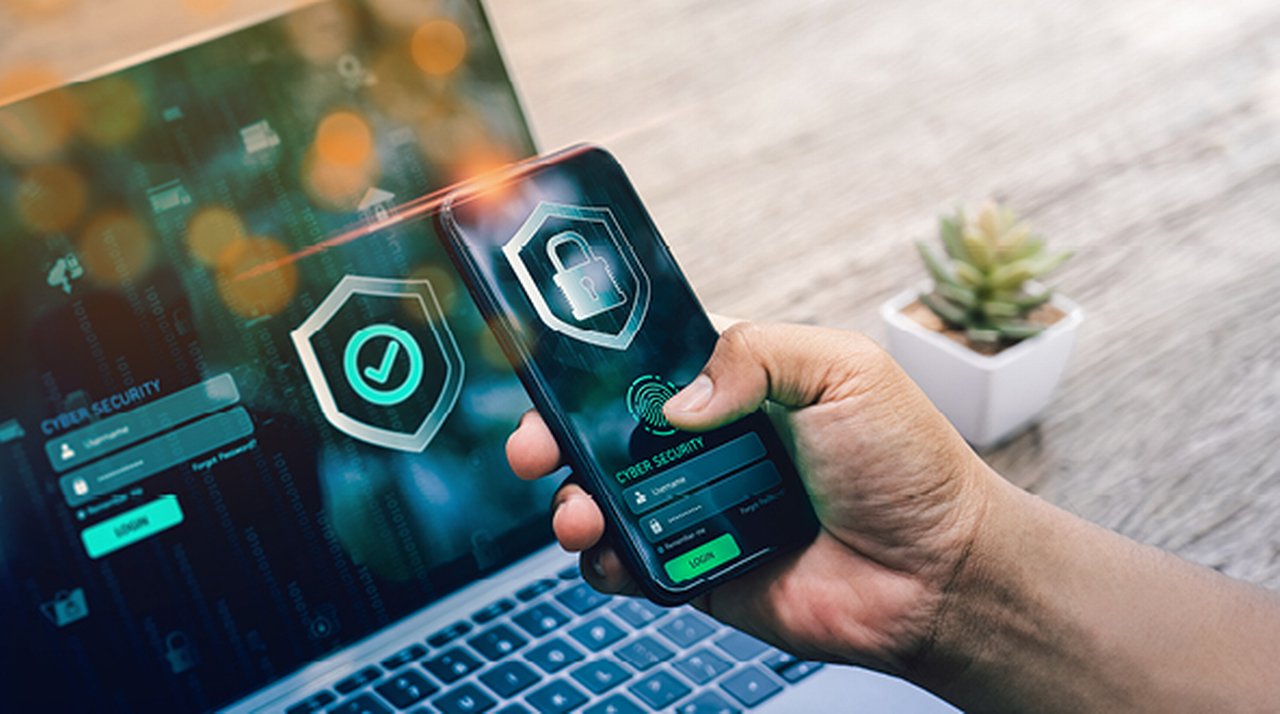 Eine Person hält ein Smartphone mit einem Cyber-Security-Login-Bildschirm, im Hintergrund ein Laptop mit ähnlicher Anzeige
