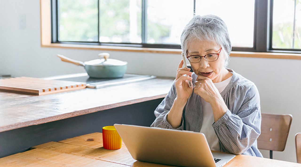 Elderly woman with a phone to her ear sits at her kitchen table with an open laptop in front of her