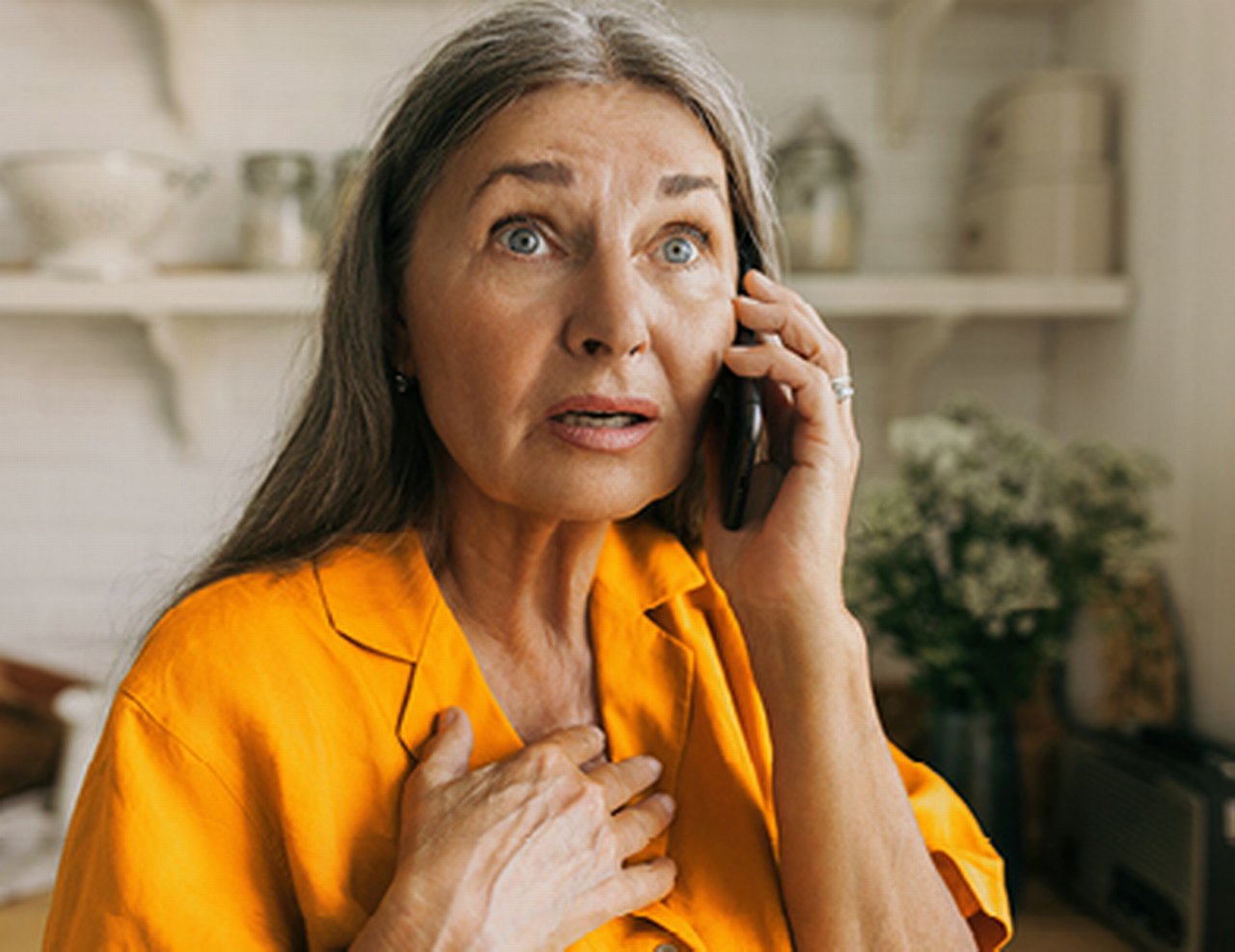 Woman with a shocked expression on her face clutches her heart with her hand while holding a phone to her ear.