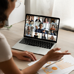 A woman sits at a desk and participates in a video conference with several people via a laptop