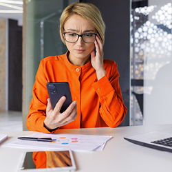 Woman sitting at a desk with a laptop and writing tools, holding a smartphone while looking at it pensively with her hand on her temple