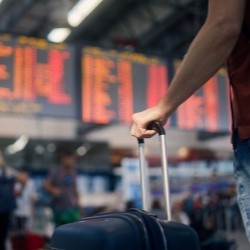 Im Vordergrund der Arm und die Hand eines Mannes, der einen Trolley h&auml;lt, im Hintergrund eine Abflugtafel