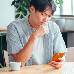 Man sitting at a table with a teacup next to him, looking thoughtfully at his smartphone with one hand on his chin